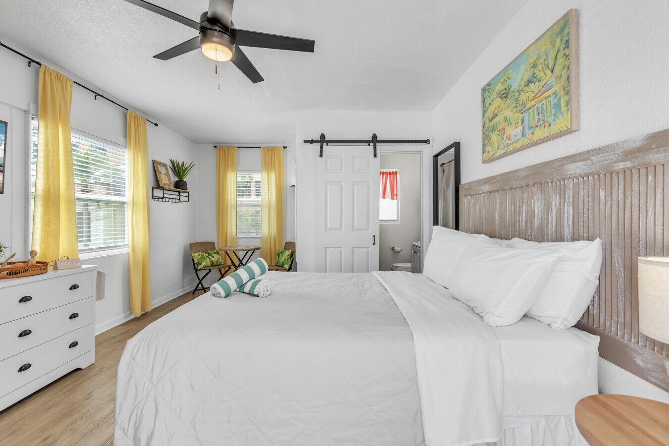 A bedroom with a bed, a ceiling fan, a window with yellow curtains, a chair, a small table, and a painting on the wall.