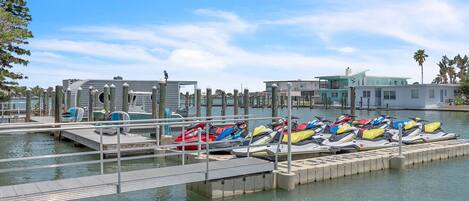 A marina with jet skis docked at wooden piers, a small dock with a boat, and buildings in the background.