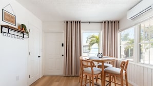 Ein Zimmer mit Holzboden, einem runden Tisch mit Stühlen, einer weißen Tür, beige farbenen Vorhängen und einer Klimaanlage.