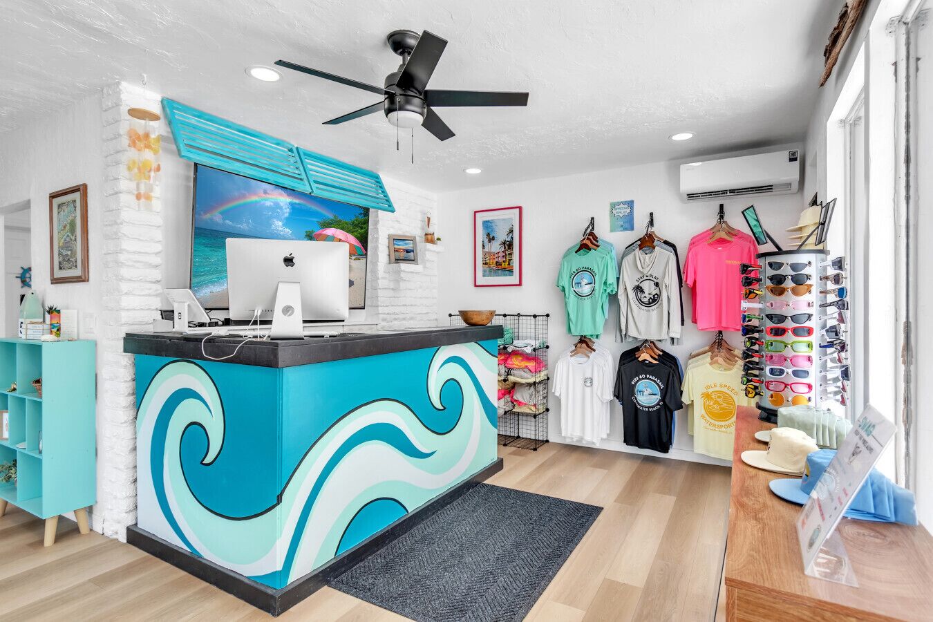 Una zona de recepción moderna con un mostrador con motivos de olas turquesas, un iMac de Apple y una variedad de ropa y gafas de sol a la vista.
