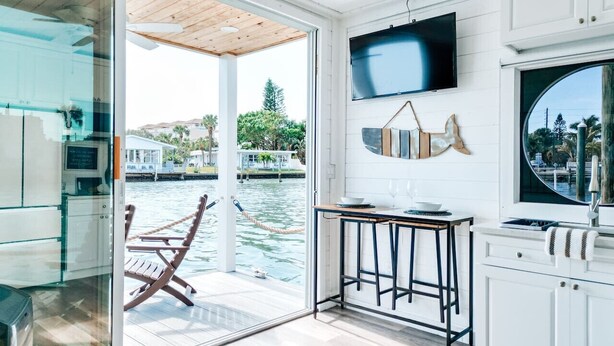 A modern kitchen with a large TV, a dining area with a table and chairs, and a view of a water body outside.