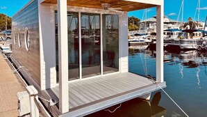 A modern floating house with large glass windows, a wooden deck, and a marina setting in the background.