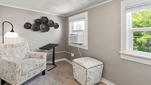 Una habitación con un sillón estampado, una mesita, un adorno de pared y una ventana con un equipo de aire acondicionado.