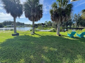A grassy area with two hammocks, two turquoise Adirondack chairs, and a body of water in the background.