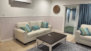 Een woonkamer met een beige bank, een donkere salontafel, vloerkleden met een patroon en een airconditioner aan de muur.