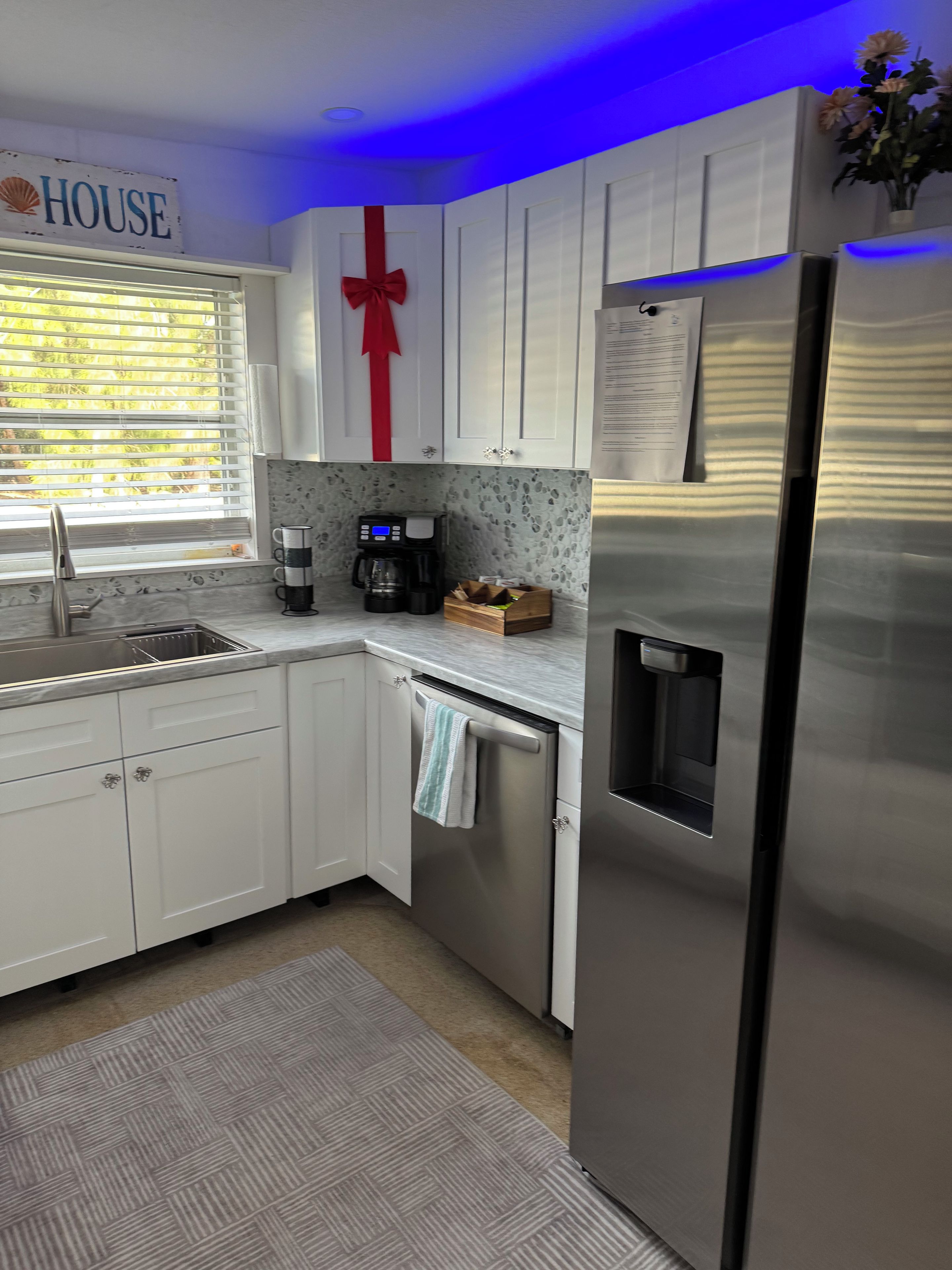 A modern kitchen with white cabinets, a stainless steel refrigerator, a coffee maker, and a window with blinds.