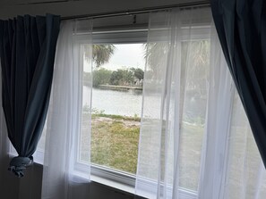 A window with sheer curtains, a view of a body of water, and a grassy area. - Resting Place of Treasure Island NPR Waterfront Retreat w/ Kayaks & Sunroom (New Port Richey)