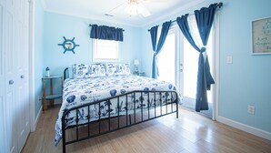 A bedroom with a bed, a ceiling fan, a window with curtains, a small table with a lamp, and a wall decoration.
