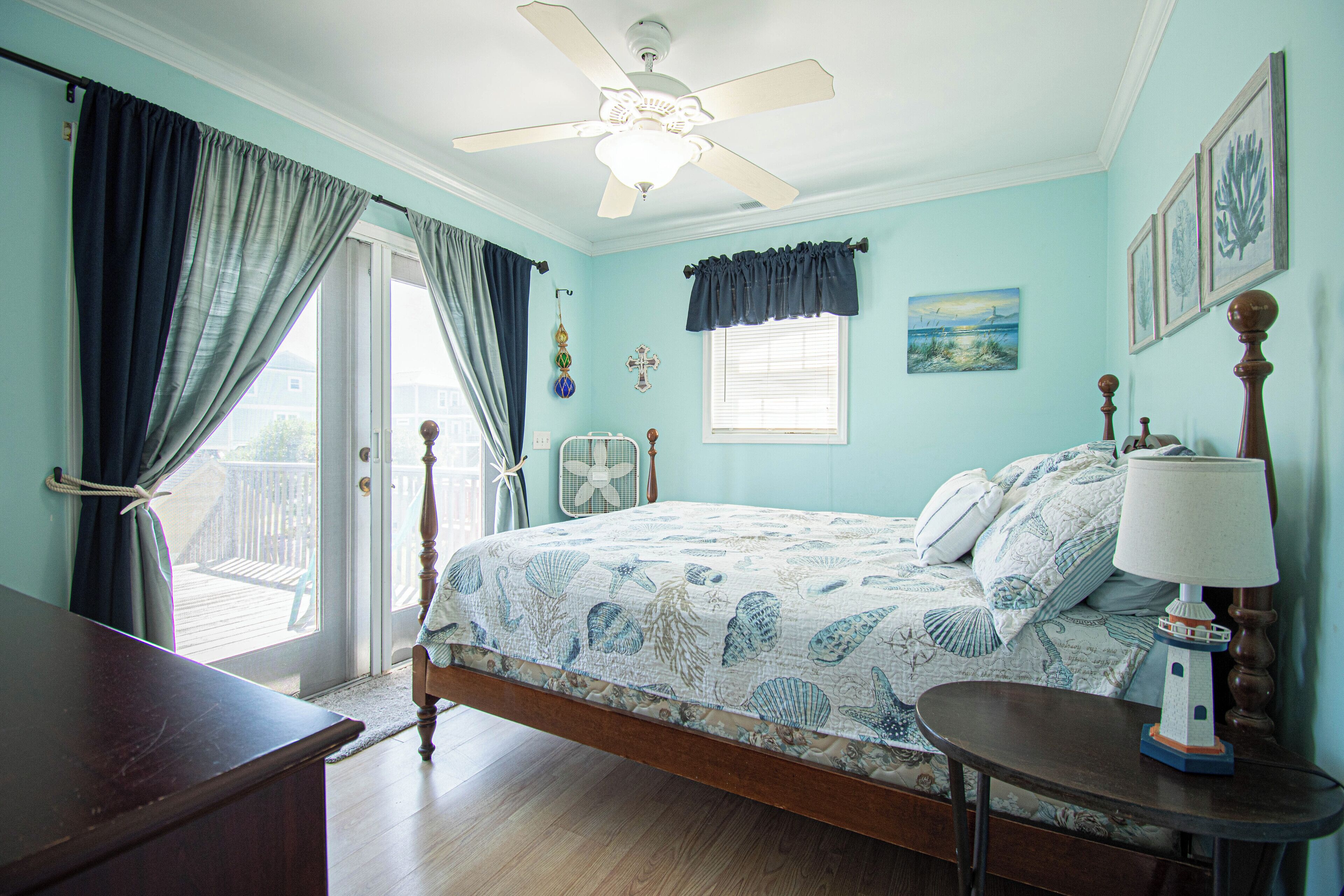 A bedroom with a bed, a ceiling fan, a window with curtains, a bedside table with a lamp, and a small fan on the floor.