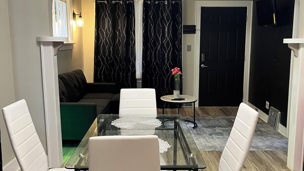 Ein Essbereich mit einem Glastisch, weißen Stühlen, einem grünen Sofa und einem schwarzen Vorhang im Hintergrund.