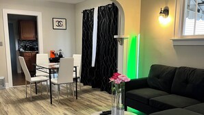 Ein Wohnzimmer mit schwarzem Sofa, einem runden Couchtisch mit Blumenvase, einem Essbereich mit Stühlen und Tisch sowie einer Küche im Hintergrund.