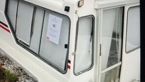 Un bateau à quai, immobile, doté d’une cabine, de fenêtres et d’une porte, amarré près d’une étendue d’eau, avec une zone herbeuse et des arbres en arrière-plan.