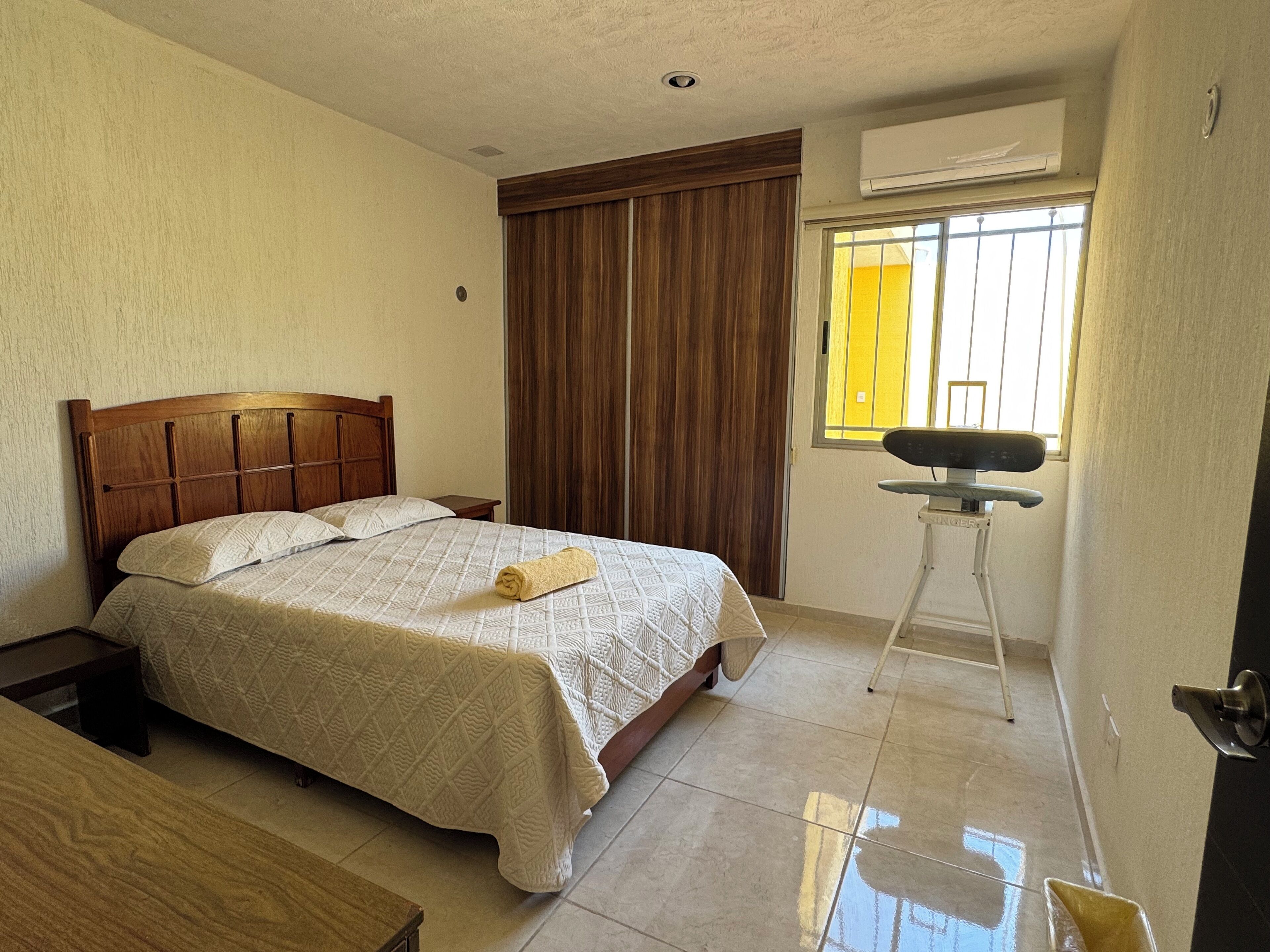 A bedroom with a wooden bed, a bedside table, a window with blinds, and an air conditioning unit.