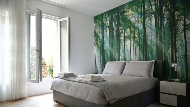 A bedroom with a bed, a nightstand, a window with curtains, and a forest-themed wall mural.
