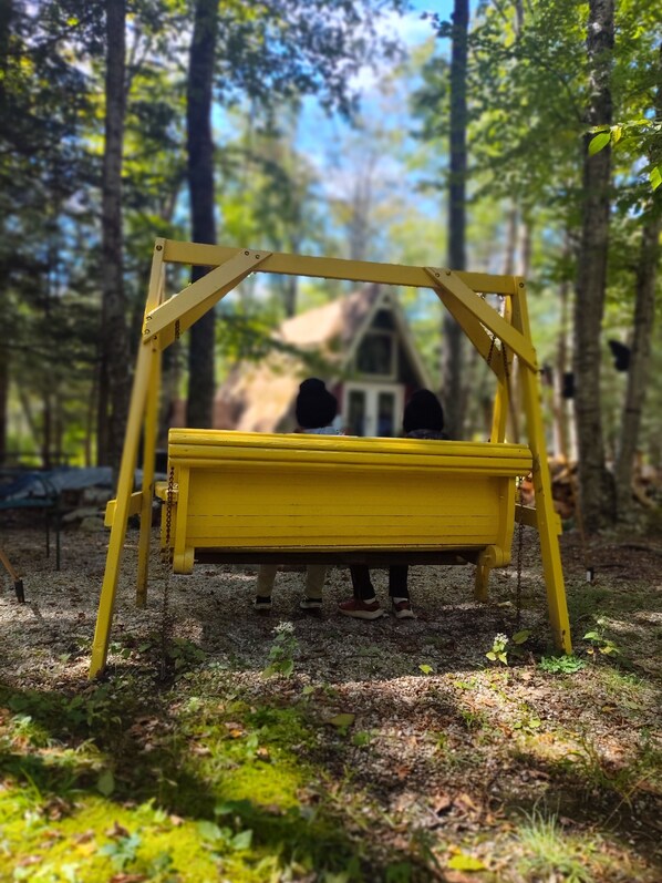 Une balançoire jaune installée dans une zone boisée, avec une petite maison en arrière-plan.