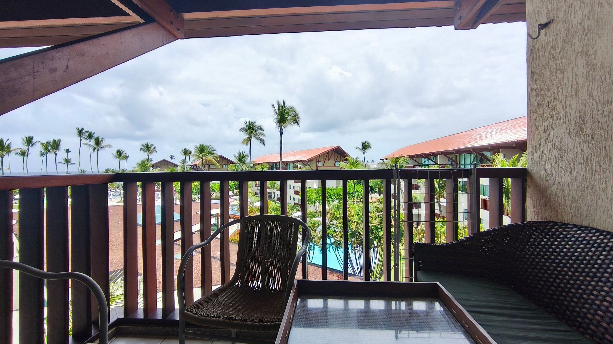A balcony with a wicker chair and a small table, overlooking a pool and palm trees.