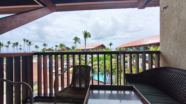 Un balcon avec un fauteuil en osier et une petite table, donnant sur une piscine et des palmiers.