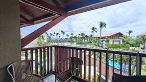 A balcony with wicker chairs, a pool, and resort buildings in the background. - Marulhos Suítes Resort (Ipojuca)