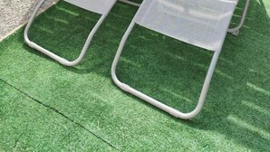 Two metal-framed lounge chairs with mesh backs and feet, placed on artificial grass in a garden area with a wooden fence and a potted plant in the background.