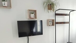 A small, modern room with a white table and two white chairs, a flat-screen TV, a hanging plant, and a wooden shelf with a clock.