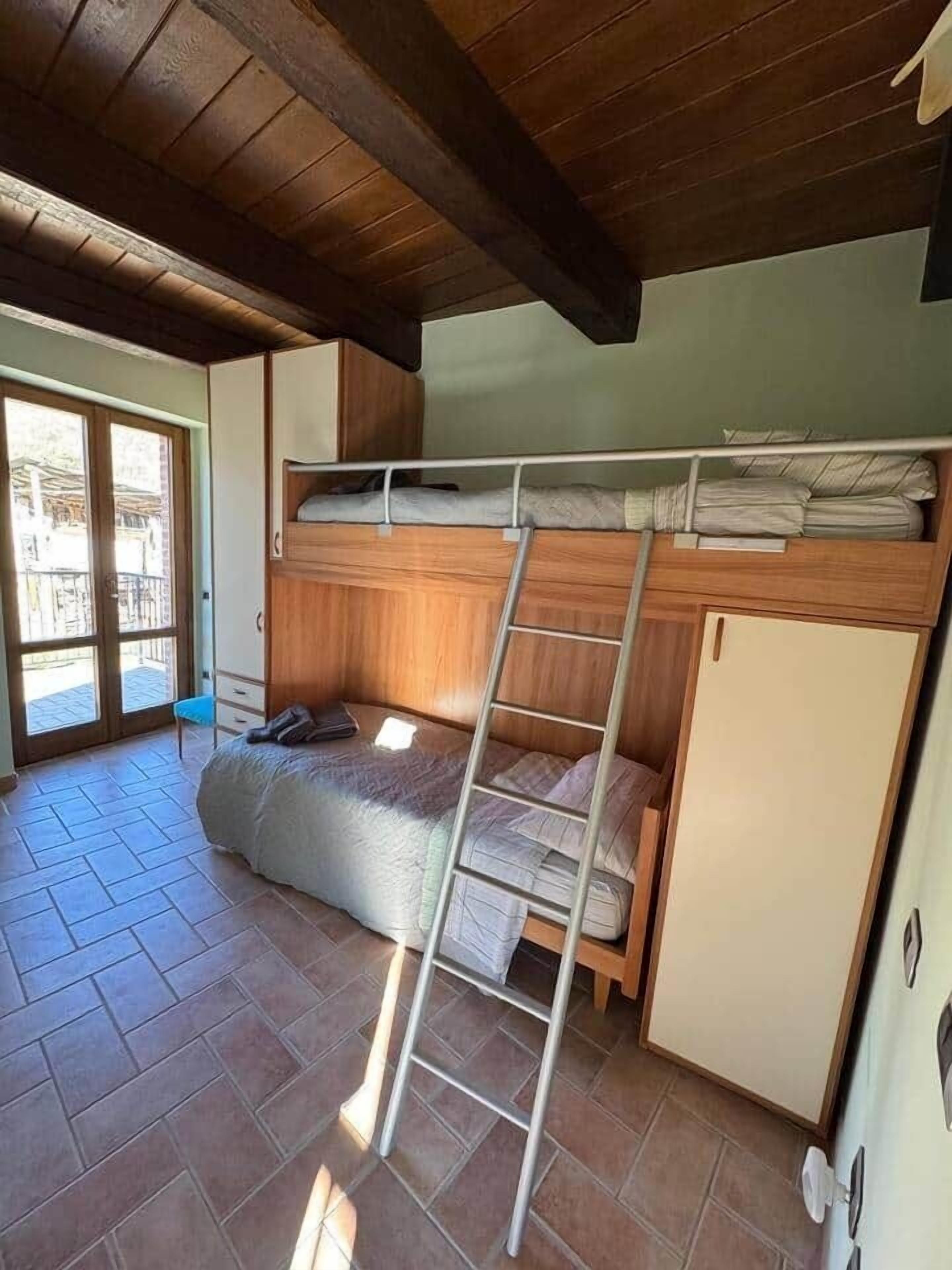 A compact room with a bunk bed, a ladder, a small refrigerator, and a sliding glass door leading to an outdoor area.