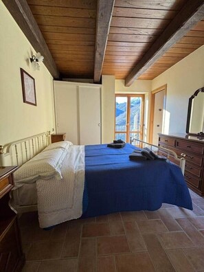 A bedroom with a bed, wooden ceiling beams, a dresser, and a door leading to a balcony with a mountain view. - Villa Paterno (Marche)