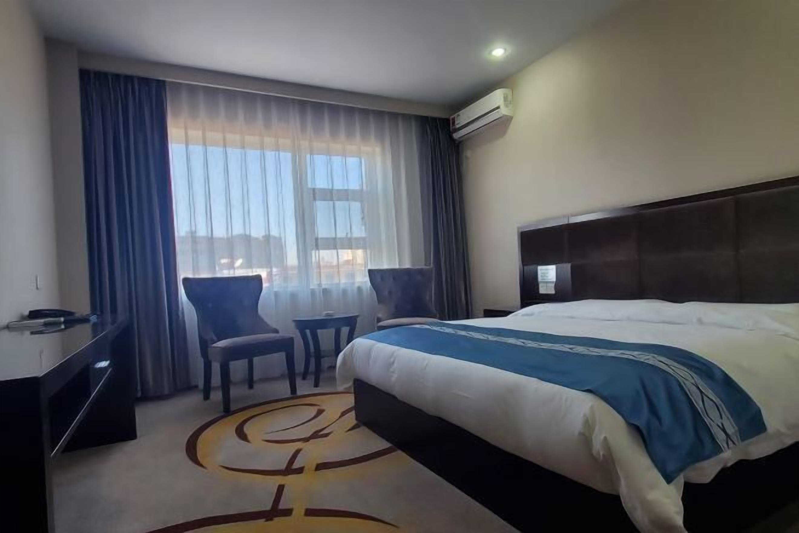 A hotel room with a bed, a desk, two chairs, a window with curtains, and an air conditioning unit.