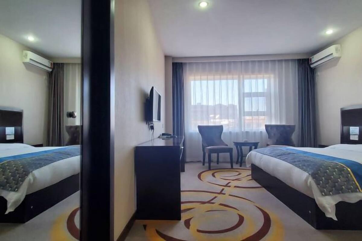 A hotel room with two beds, a desk, a chair, and a view of the city through the window.