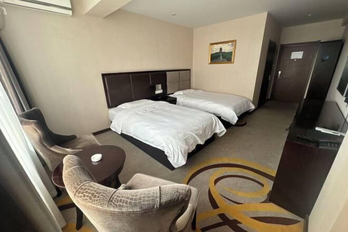 A hotel room with two beds, a small table, a chair, a painting on the wall, and a door.