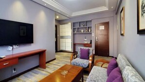 Una sala de estar con una televisión de pantalla plana, una mesa de centro de madera, un sofá y una silla con un cojín estampado.