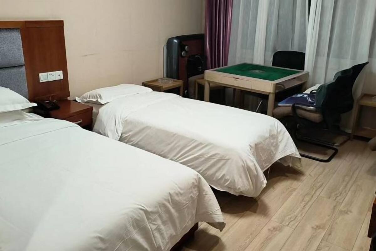 A hotel room with two beds, a desk with a green table, a chair, a small table, and a wall socket.
