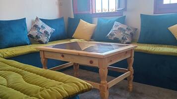 Un salon chaleureux avec une table basse en bois, des canapés bleus et verts, et une fenêtre ornée de barreaux décoratifs.