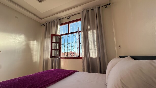 Une chambre à coucher avec un lit, une couverture violette, une fenêtre avec des barreaux en métal et des rideaux.