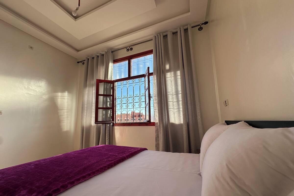 Une chambre à coucher avec un lit, une couverture violette, une fenêtre avec des barreaux en métal et des rideaux.