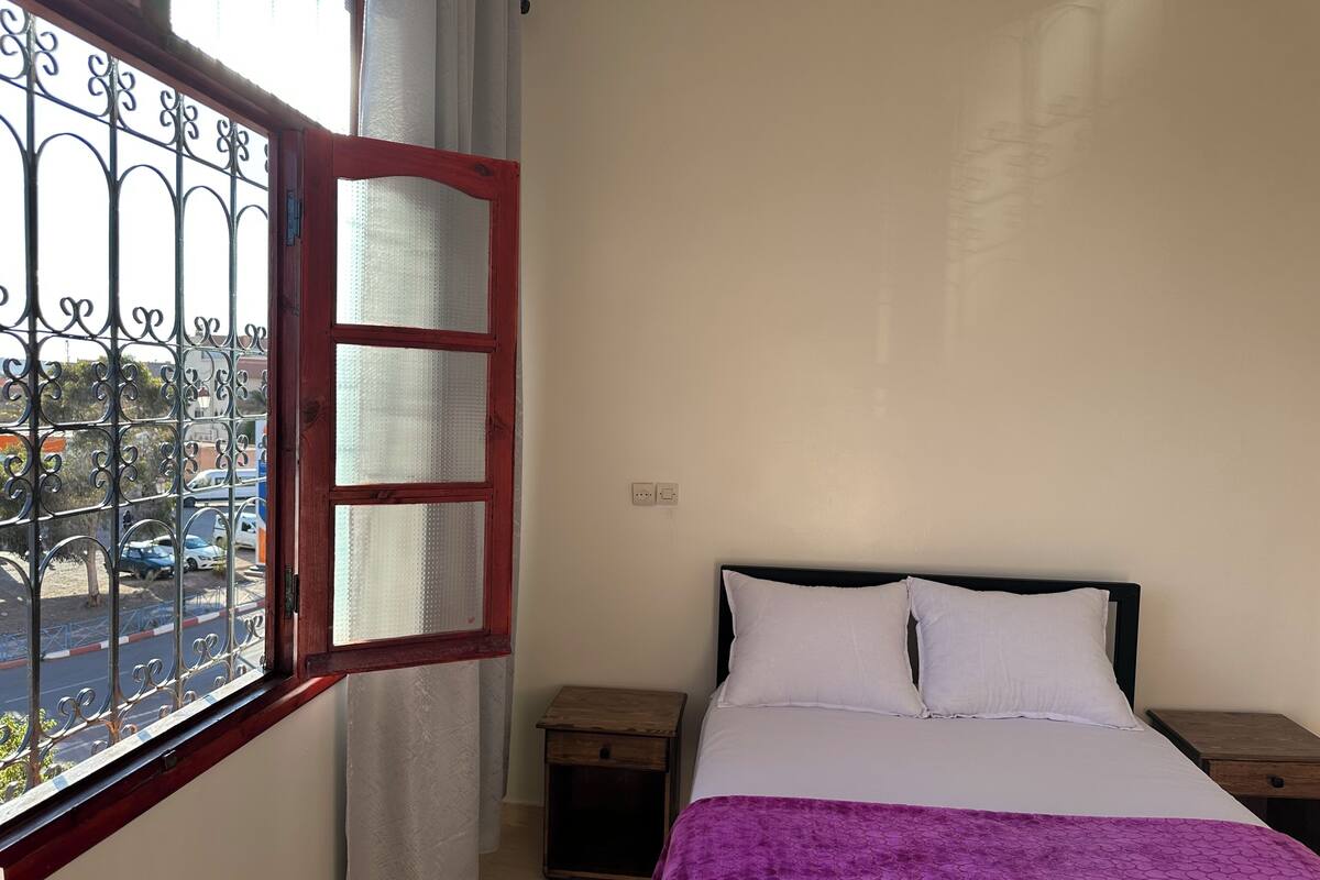 Une chambre à coucher avec un lit, une table de chevet, une commode en bois et une fenêtre avec des barreaux en métal.