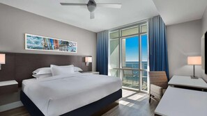 Une chambre d’hôtel avec un grand lit, un ventilateur de plafond, un tableau, une vue sur l’océan et un balcon avec une chaise.