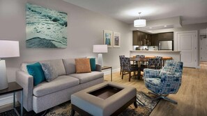 Un saló modern amb un sofà gris, una taula de centre i un butac armchair amb un disseny estampat. Hi ha una zona de menjador amb una taula i cadires, i una cuina al fons.