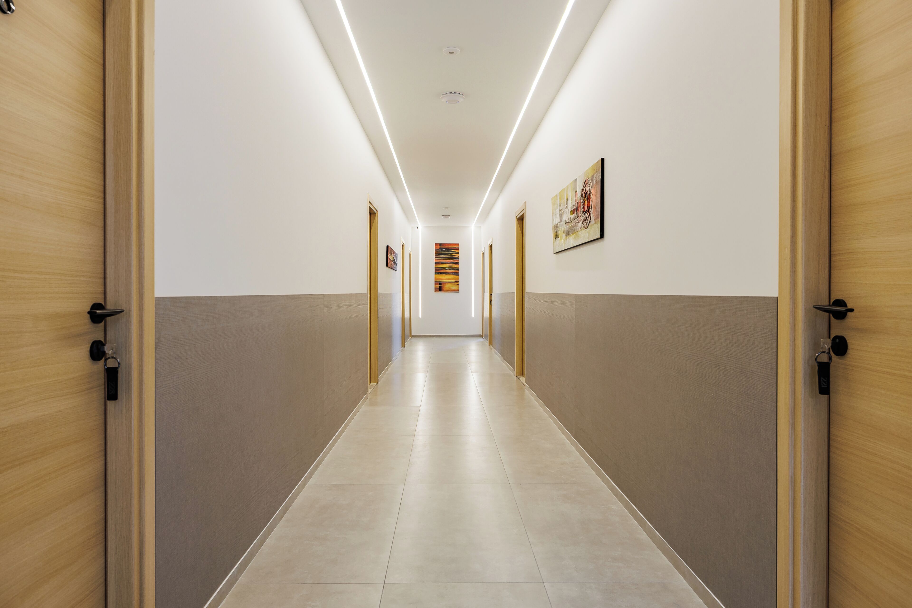 A long, narrow hallway with wooden doors on both sides, a tiled floor, and a ceiling with linear lighting.