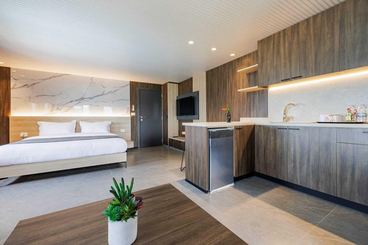 A modern hotel room with a bed, a small kitchenette, a TV, and a marble wall.