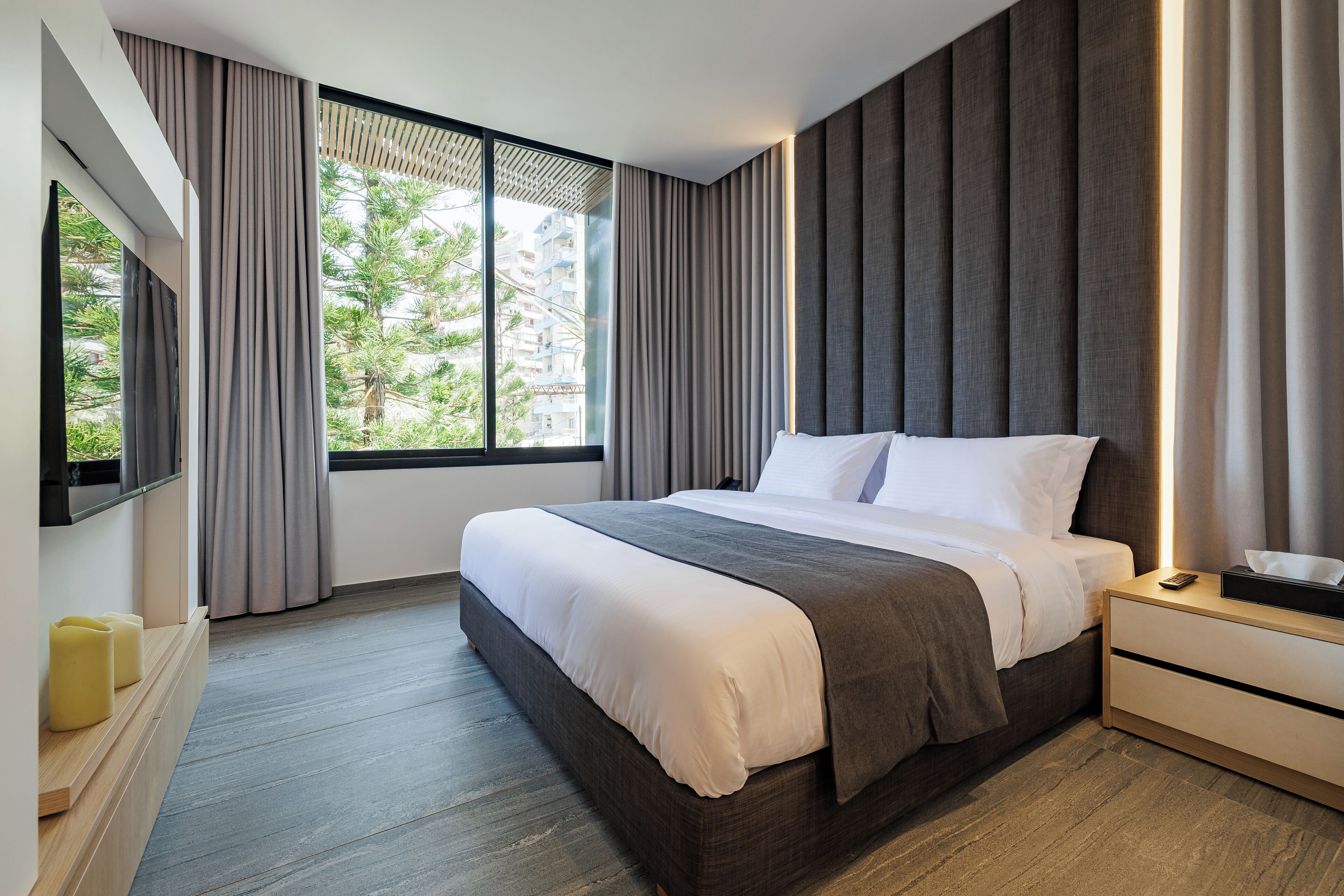 A modern hotel room with a large bed, a bedside table with a lamp, and a view of greenery outside.
