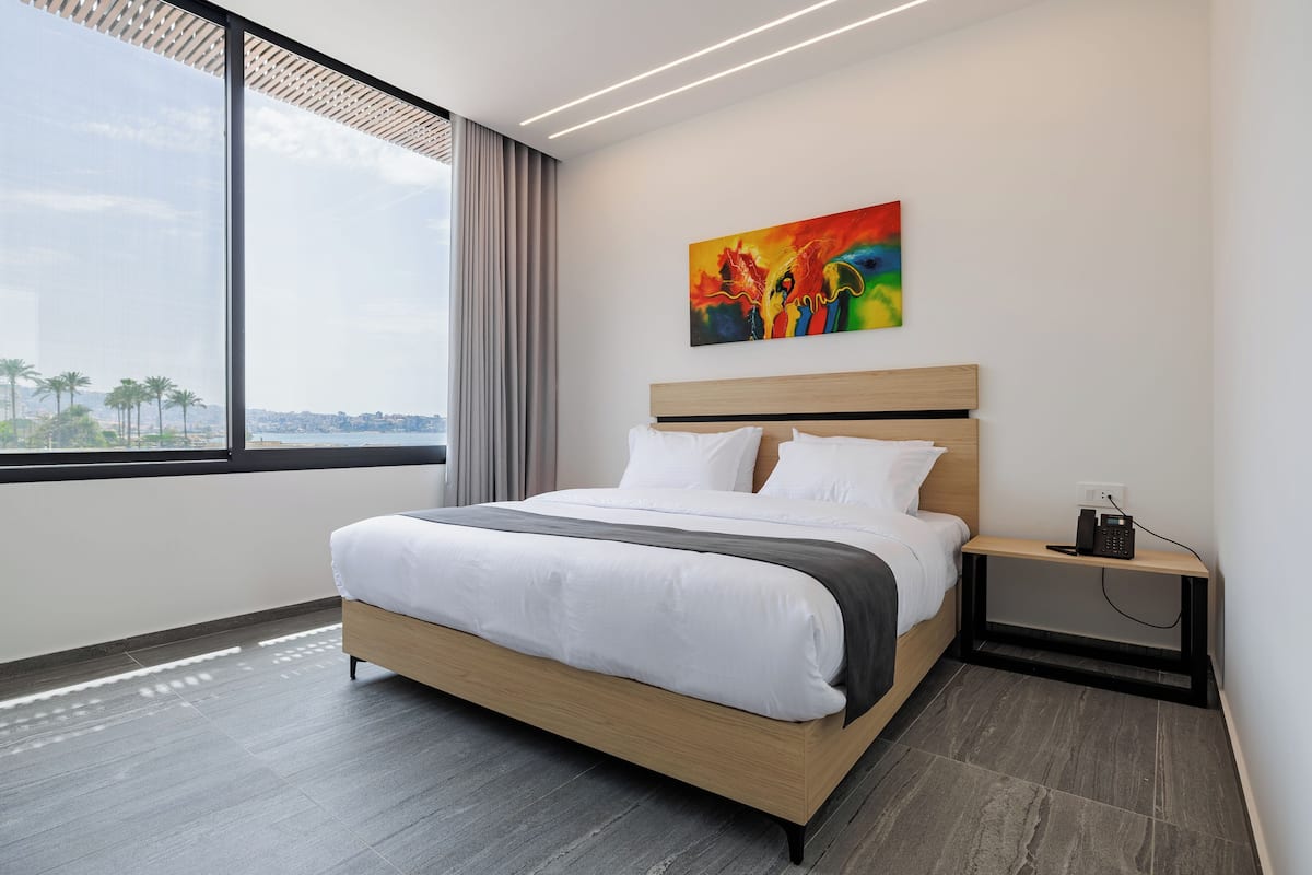 A modern hotel room with a large bed, a nightstand, a painting, and a view of palm trees.
