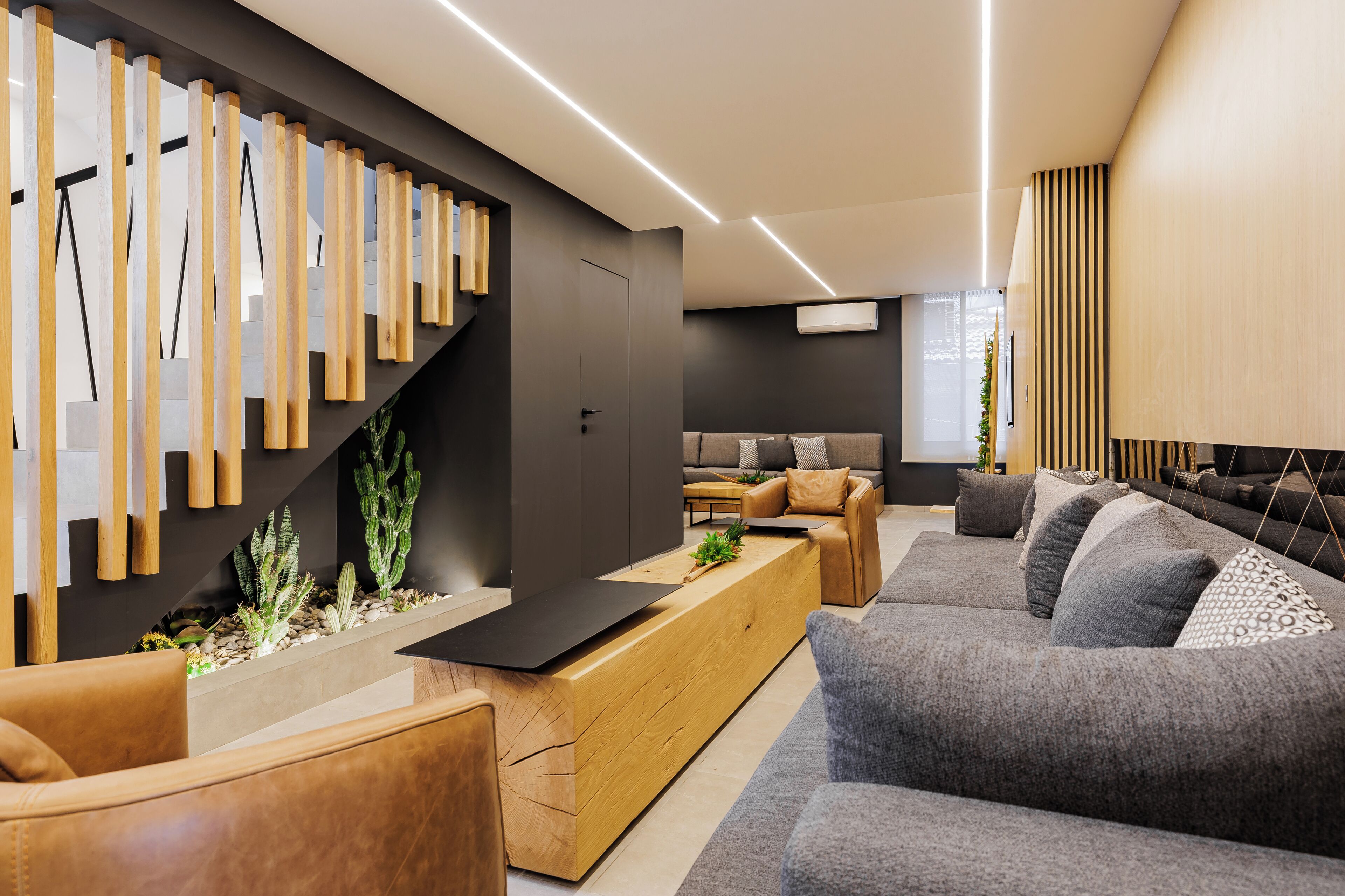 A modern living room with a wooden reception desk, grey sofas, and a staircase with wooden railings.
