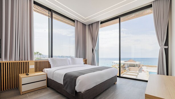 Un dormitorio moderno con una cama grande, mesitas de noche de madera y un escritorio. Cuenta con ventanas amplias que ofrecen vistas al mar y al paisaje urbano.