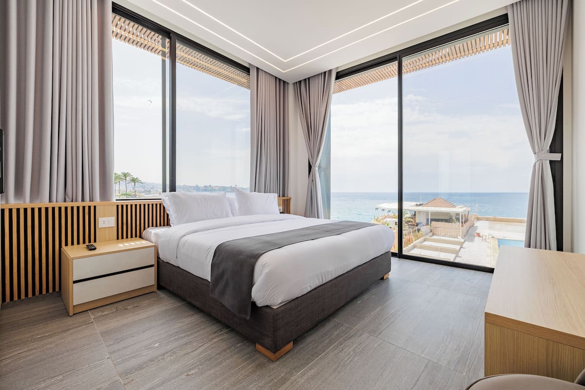 A modern bedroom with a large bed, wooden nightstands, and a desk. It features large windows with a view of the sea and a cityscape.