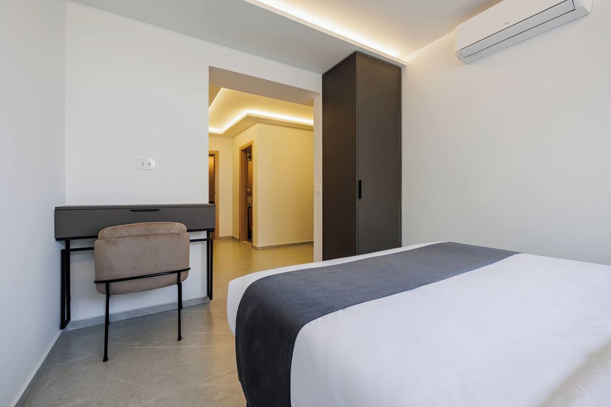 A hotel room with a bed, a desk, and a chair. There is a wardrobe and an air conditioning unit on the wall.