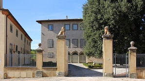 Un edificio con entrada protegida por un portón, un camino de entrada que lleva a la puerta principal, y un gran árbol al fondo.