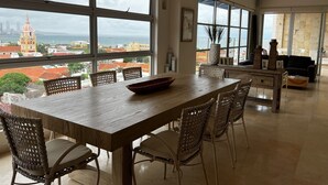 Un comedor con mesa de madera y sillas de mimbre, ventanas amplias con vista a la ciudad y ventiladores de techo.