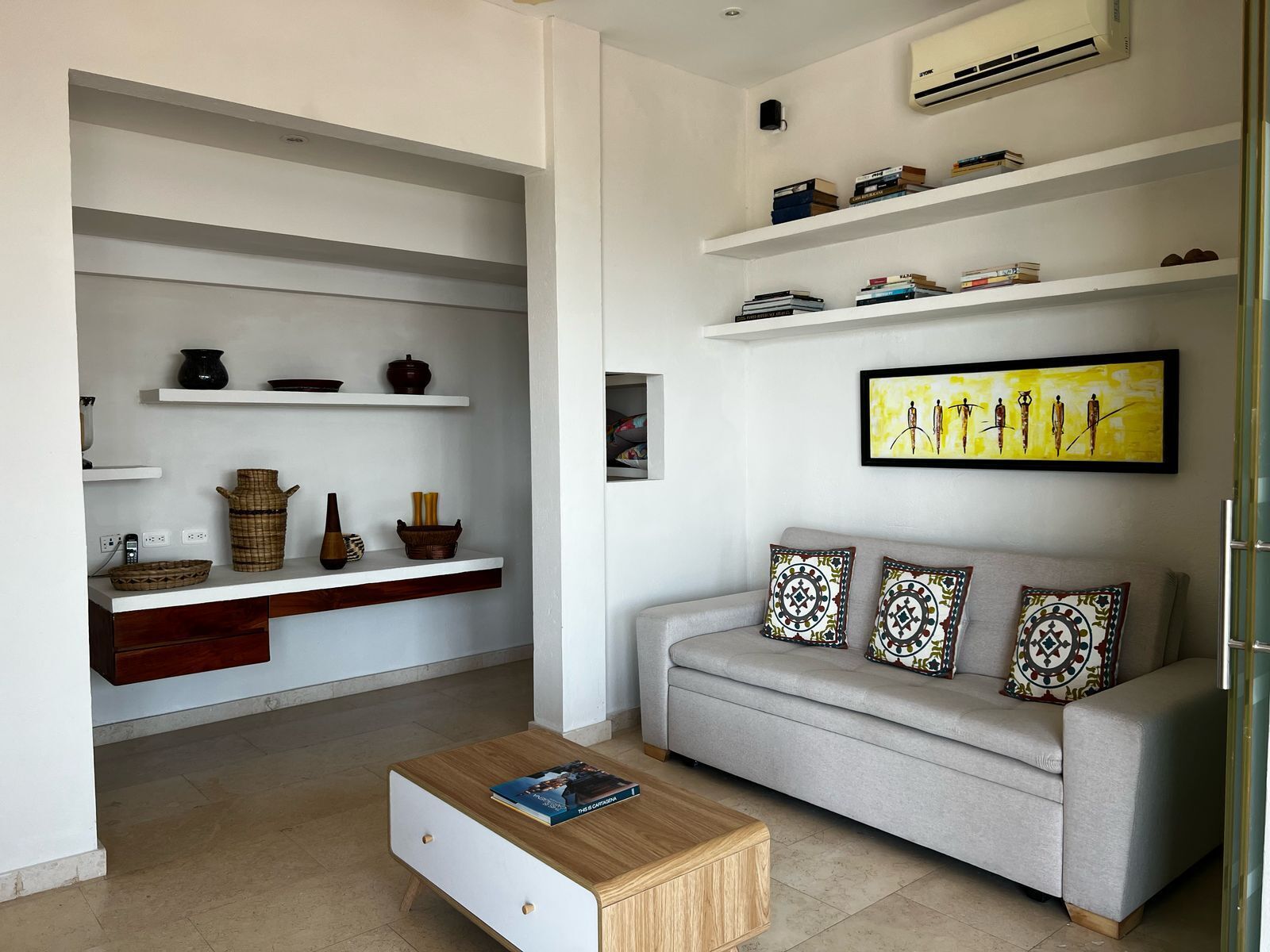 A modern living room with a grey sofa, patterned cushions, a coffee table with a book, and shelves with decorative items and books.