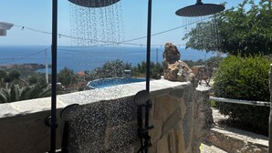 A showerhead with water flowing over a stone ledge, overlooking a pool and the sea.