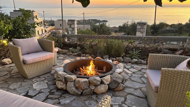 A patio with wicker furniture, a stone fire pit, and a view of the sea at sunset.